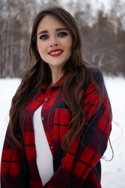 Karlı kış ormanlarında dikilip kameraya bakarken kırmızı dudaklı, karlı gömlekli, saçlarına dokunan çekici bir kadın.