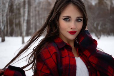 Karlı kış ormanlarında dikilip kameraya bakarken kırmızı dudaklı, karlı gömlekli, saçlarına dokunan çekici bir kadın.