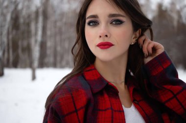 Karlı kış ormanlarında dikilip kameraya bakarken kırmızı dudaklı, karlı gömlekli, saçlarına dokunan çekici bir kadın.