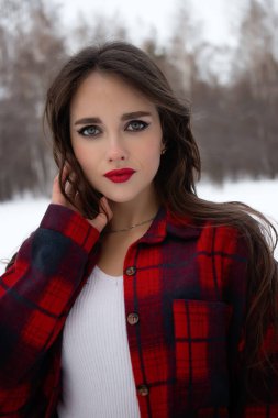 Karlı kış ormanlarında dikilip kameraya bakarken kırmızı dudaklı, karlı gömlekli, saçlarına dokunan çekici bir kadın.