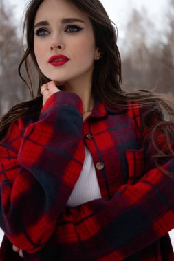 Karlı kış ormanlarında dikilip kameraya bakarken kırmızı dudaklı, karlı gömlekli, saçlarına dokunan çekici bir kadın.