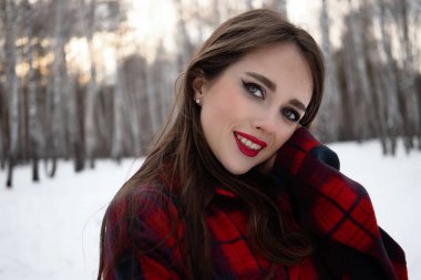 Karlı kış ormanlarında dikilip kameraya bakarken kırmızı dudaklı, karlı gömlekli, saçlarına dokunan çekici bir kadın.