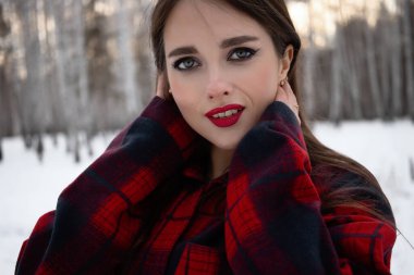 Karlı kış ormanlarında dikilip kameraya bakarken kırmızı dudaklı, karlı gömlekli, saçlarına dokunan çekici bir kadın.