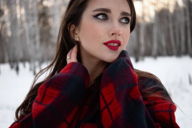 Karlı kış ormanlarında dikilip kameraya bakarken kırmızı dudaklı, karlı gömlekli, saçlarına dokunan çekici bir kadın.