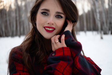 Karlı kış ormanlarında dikilip kameraya bakarken kırmızı dudaklı, karlı gömlekli, saçlarına dokunan çekici bir kadın.