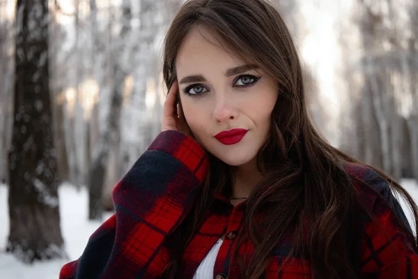 Karlı kış ormanlarında dikilip kameraya bakarken kırmızı dudaklı, karlı gömlekli, saçlarına dokunan çekici bir kadın.