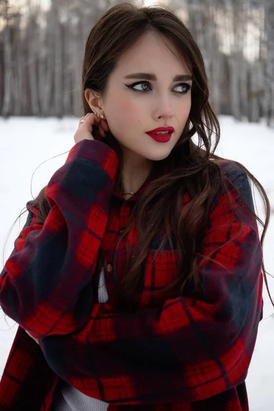 Karlı kış ormanlarında dikilip kameraya bakarken kırmızı dudaklı, karlı gömlekli, saçlarına dokunan çekici bir kadın.