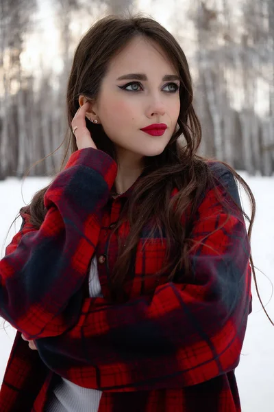 Karlı kış ormanlarında dikilip kameraya bakarken kırmızı dudaklı, karlı gömlekli, saçlarına dokunan çekici bir kadın.
