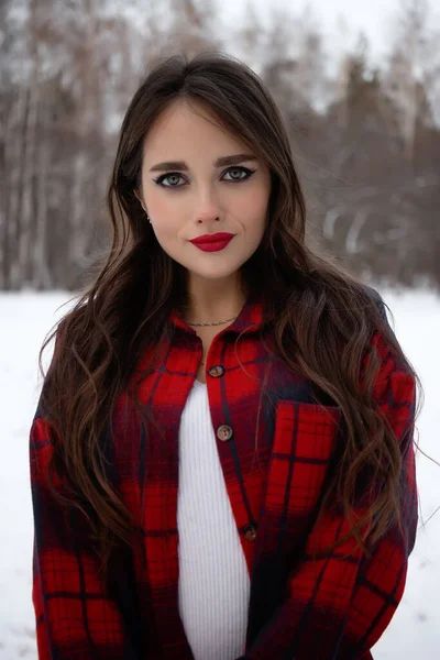 Karlı kış ormanlarında dikilip kameraya bakarken kırmızı dudaklı, karlı gömlekli, saçlarına dokunan çekici bir kadın.