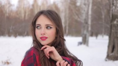 Kış ormanında dalgalı saçlı genç bir kadın ayakta duruyor ve yüzüne dokunuyor.