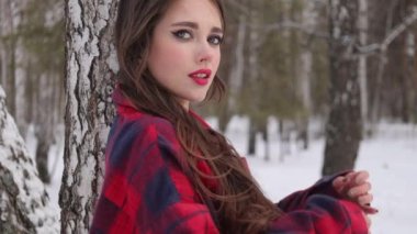 Kış ormanında dalgalı saçlı genç bir kadın ayakta duruyor ve yüzüne dokunuyor.