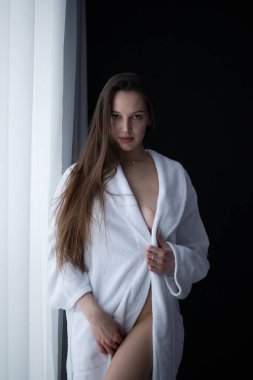 Çıplak göğüslü, odanın penceresinin yanında duran ve kameraya bakan seksi kadının alçak açılı yan görüntüsü.