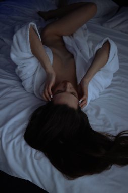 Üstsüz, bornozlu seksi bir kadın yatak odasında rahat bir yatakta yatıyor.