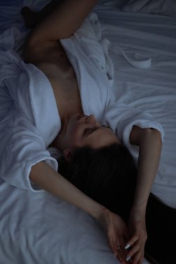 Üstsüz, bornozlu seksi bir kadın yatak odasında rahat bir yatakta yatıyor.