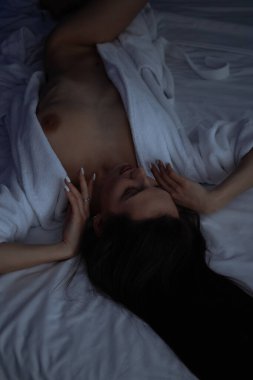 Üstsüz, bornozlu seksi bir kadın yatak odasında rahat bir yatakta yatıyor.