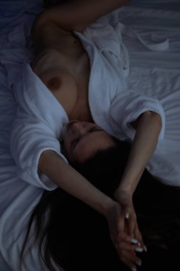 Üstsüz, bornozlu seksi bir kadın yatak odasında rahat bir yatakta yatıyor.