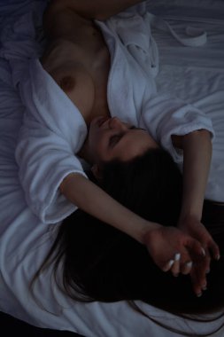 Üstsüz, bornozlu seksi bir kadın yatak odasında rahat bir yatakta yatıyor.