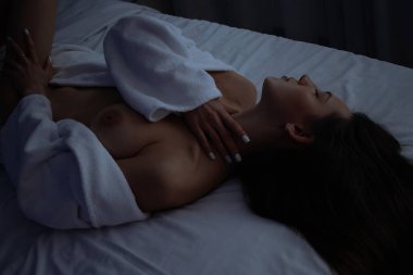 Üstsüz, bornozlu seksi bir kadın yatak odasında rahat bir yatakta yatıyor.