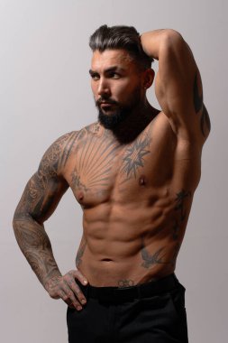 Hispanic shirtless male model with muscular tattooed torso standing with hands in pockets and looking away on gray backdrop clipart