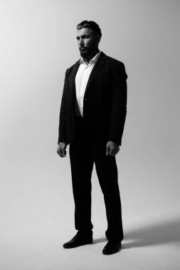 Siyah yelekli, beyaz gömlekli, sakallı adam stüdyoda elleri ceplerinde kameraya bakıyor.