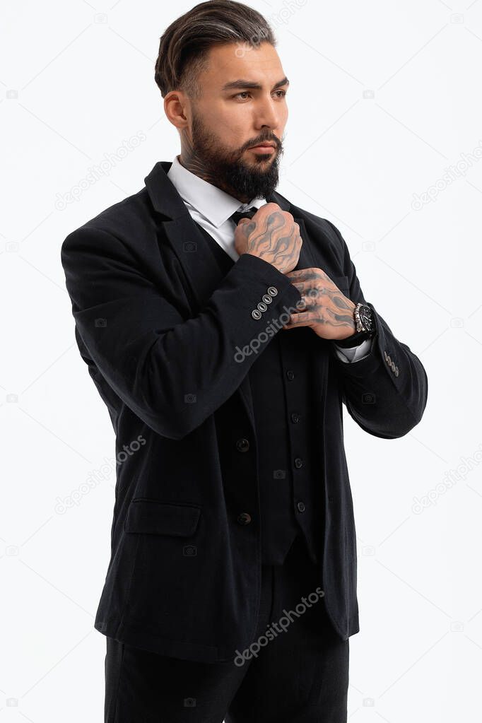 Confident Hispanic bearded man with tattooed hands in white shirt and black trousers adjusting tie and looking away while standing against gray background
