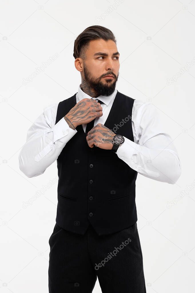 Confident Hispanic bearded man with tattooed hands in white shirt and black trousers adjusting tie and looking away while standing against gray background