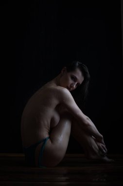 Sıska, çıplak ayaklı, siyah saçlı, bikinili, sutyensiz, duşta tahta zeminde oturan ve çıplak göğsünü kapatan genç bir kadın.