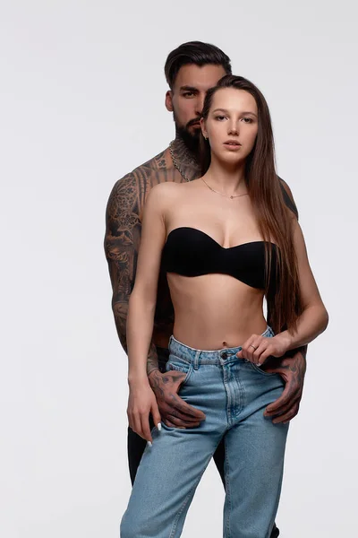 Kaslı, dövmeli, gri arka planda külotlu, zayıf, üstsüz kadını kucaklayan adam.