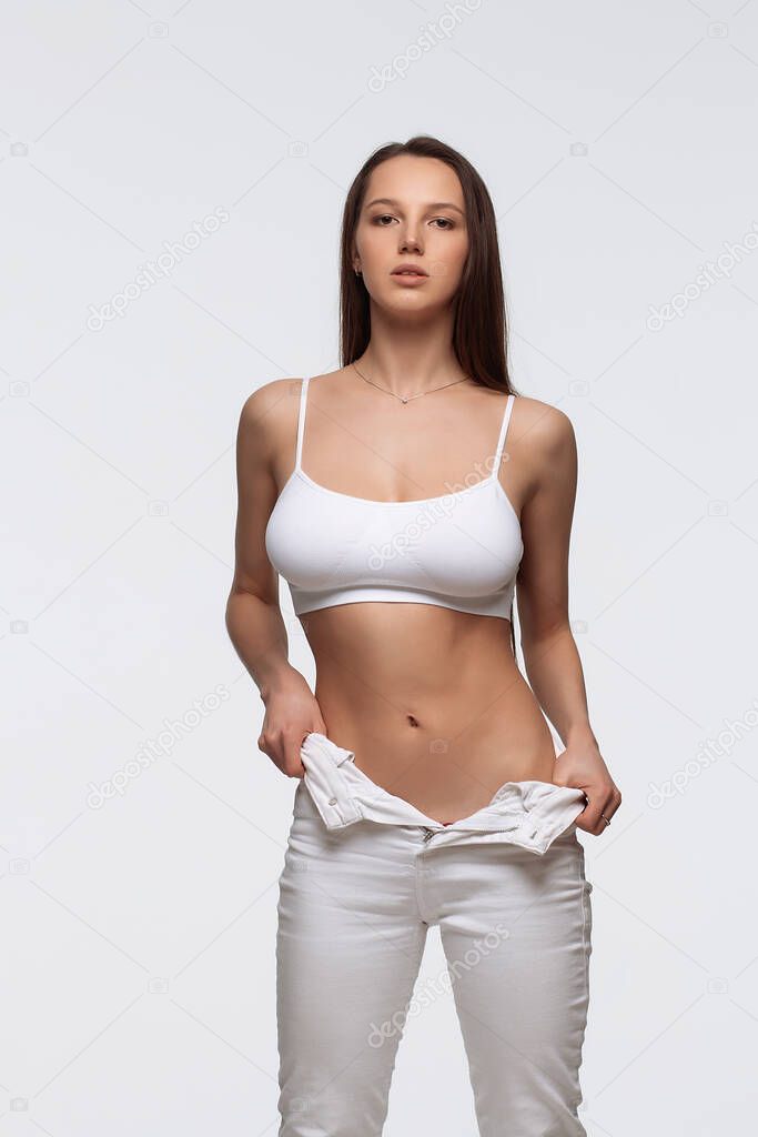 Self assured young female model with long straight dark hair in white bra and jeans standing in light studio with hands in pockets and looking at camera