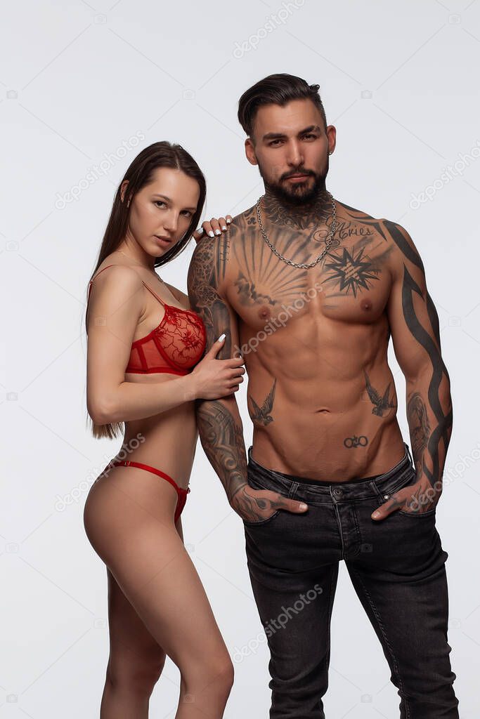 Confident tattooed bearded male standing with seductive female partner wearing red lace lingerie in studio and looking at camera