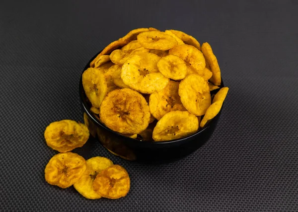Fried banana chips or banana wafers, arranged beautifully in a black ceramic bowl with a black textured background.