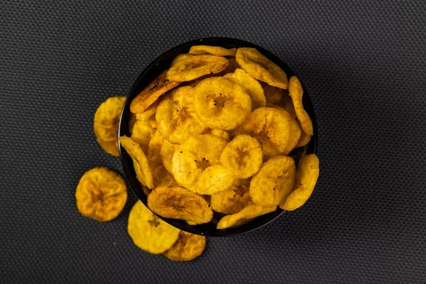 Fried banana chips or banana wafers, arranged beautifully in a black ceramic bowl with a black textured background. Top view