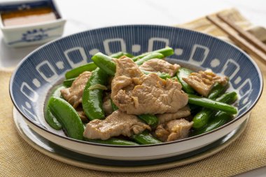 Stir fried chicken breast slices with sugar snap peas, a simple Thai Chinese dish, served on a deep blue plate with a small cup of garlic chili relish.