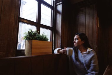 Yalnız genç bir kadın kahve içiyor ve pencereden dışarı bakıyor.