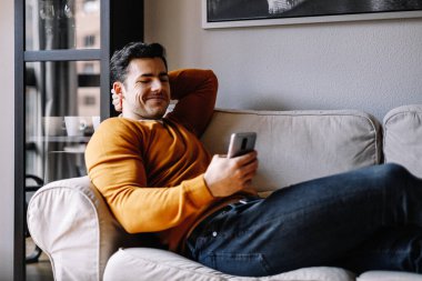 İspanyol genç adam cep telefonunu kullanarak kanepede dinleniyor.