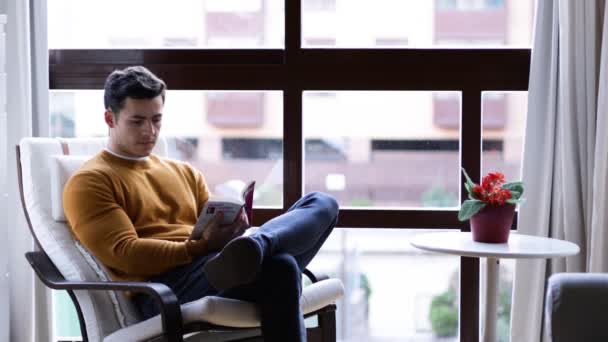 Молодий Стильний Латинський Чоловік Читає Книгу Вітальні Поруч Вікном — стокове відео