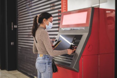 Maskeli genç bir kadın ATM makinesinden para alıyor.
