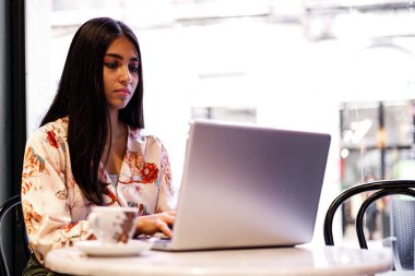 Güzel, genç ve çekici bir Hintli kadın dizüstü bilgisayarını kullanırken gülümsüyor. Parlak bir sabahta modaya uygun bir kafede oturuyor ve tek başına dinleniyor..