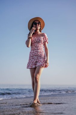 Kırmızı elbiseli, şapkalı ve güneş gözlüklü esmer kadın telefonla konuşuyor, kumsalda yürüyor.