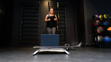 Genç bir kadın spor salonunda dizüstü bilgisayar egzersizleri yapıyor ve online dersler alıyor.