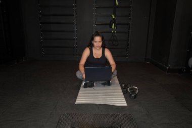 Bacaklarında dizüstü bilgisayarla spor salonunun paspasında oturan genç bir kadın halterlerle kameraya bakıyor.