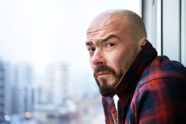 Pencerede sıradan giysiler içinde sakallı çekici kel bir adamın portresi, somurtkan, şaşkın ve hoşnutsuz bir şekilde kameraya bakıyor. Pencerenin dışında büyük bir şehrin bulanık bir manzarası var..