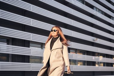 portrait of African American woman with sunglasses and beige coat walking with suitcase in hand outside station.
