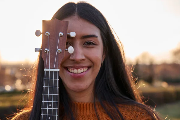Ukulele ile yüzünü örten Latin kadın. Kahverengi saçlı, gülümseyen genç bir kadın..