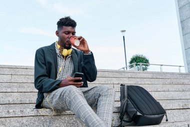 Havalı bir Afro-Amerikalı 'nın açık hava portresi kağıt bardakta kahve içen ve internet sitelerinde cep telefonunda online bir uygulama kullanarak yeni ilginç yerler arayan.