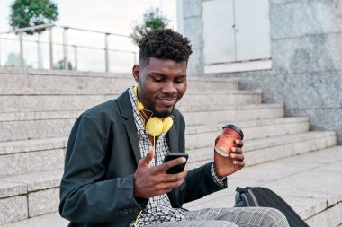 Dışarıda spor ceket giyen, elinde kahve fincanı tutan, cep telefonu kullanan neşeli Afrikalı genç bir adam.