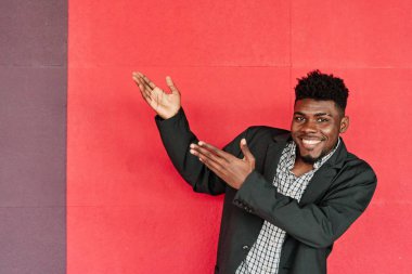 Spor ceketli adam kırmızı arkaplandaki boşluğu gösteriyor. Genç Afro-Amerikan adam gülümsüyor..