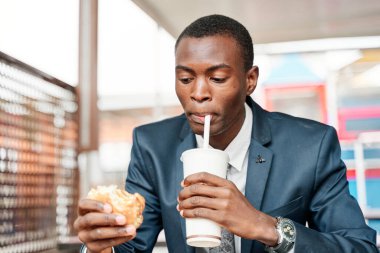 Afro-Amerikan adam elinde hamburgerle soda içiyor.