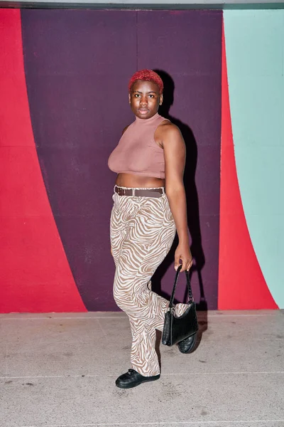 Elinde çantasıyla mor bir arka planda poz veren genç bir Afro-Amerikalı kadın. Sıradan bir kıyafet giyiyor ve kısa kızıl saçlı..