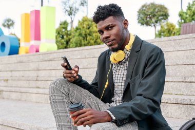 Dışarıda spor ceket giyen, elinde kahve fincanı tutan, cep telefonu kullanan neşeli Afrikalı genç bir adam.